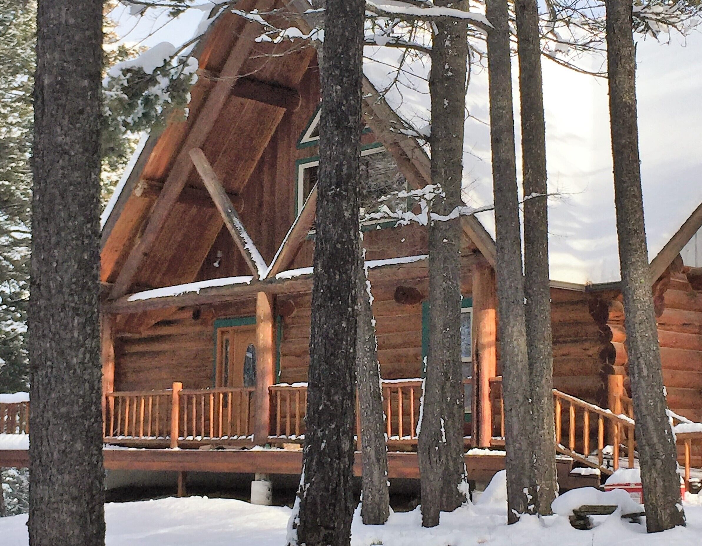 A log cabin with a wooden deck and multiple windows, surrounded by snow-covered trees.