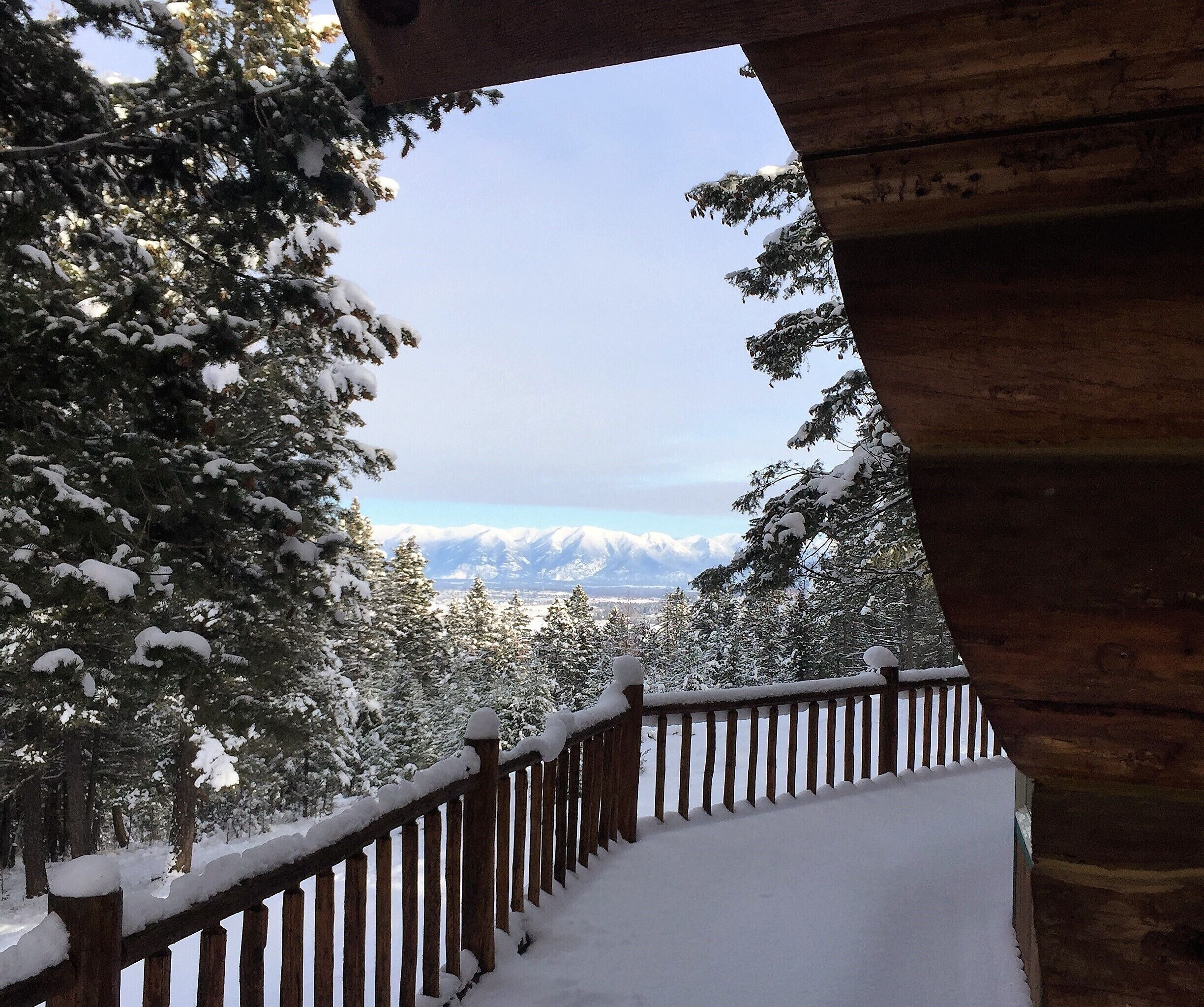 A snow-covered balcony with a wooden railing, overlooking a forest of snow-laden trees and distant mountains.