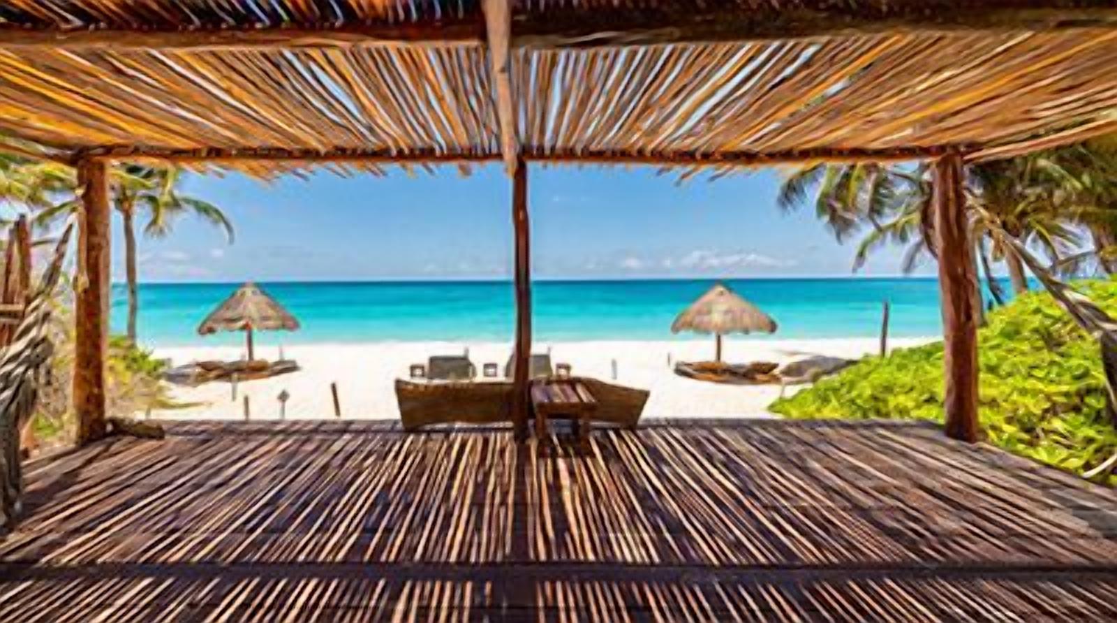A beach view from a covered area with a wooden floor, two lounge chairs, and a table.
