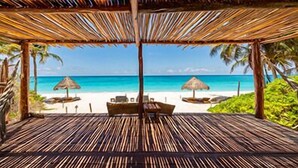 A beach view from a covered area with a wooden floor, two lounge chairs, and a table. - Casa Carolina Tulum (Tulum)