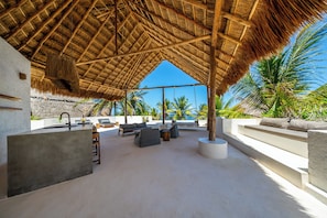 A covered outdoor seating area with a thatched roof, lounge chairs, and a view of the ocean.