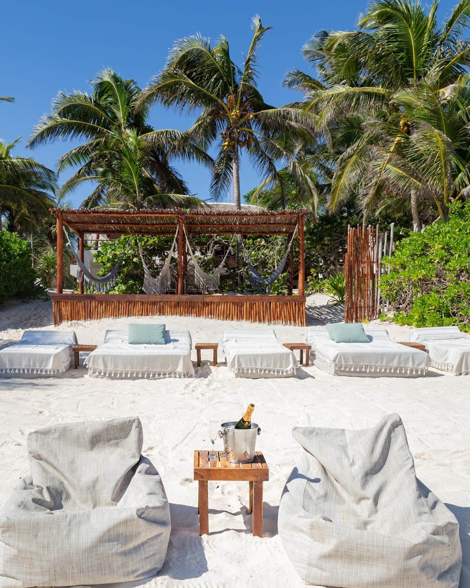 A beachside lounge with white cushioned seating, a wooden table with a bottle of champagne, and a thatched roof structure.