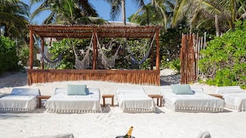 A beachside lounge with white cushioned seating, a wooden table with a bottle of champagne, and a thatched roof structure.