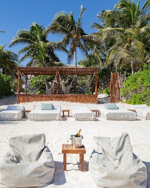 A beachside lounge with white cushioned seating, a wooden table with a bottle of champagne, and a thatched roof structure.
