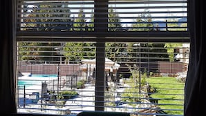Blick durch ein Fenster mit Rollläden, der einen Garten mit Swimmingpool, Grillplatz und umzäunter Rasenfläche zeigt.