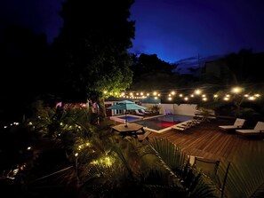 Ein Poolbereich mit Liegestühlen und Sonnenschirmen, umgeben von Lichterketten und tropischen Pflanzen.