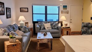 A cozy living room with a sofa, a small table, and a bed. There are wall decorations and a window with blinds. - Right on the Gulf! Beautiful white sandy beachfront! Safe, quiet, and friendly! (Treasure Island)