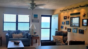 A living room with a sofa, a coffee table, and a chair. There is a window with blinds, a ceiling fan, and a door with a number on it. - Right on the Gulf! Beautiful white sandy beachfront! Safe, quiet, and friendly! (Treasure Island)