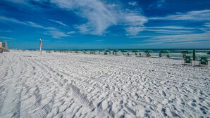 Une plage de sable fin, avec des parasols verts et des chaises longues, un ciel dégagé et une vue lointaine sur l’océan.