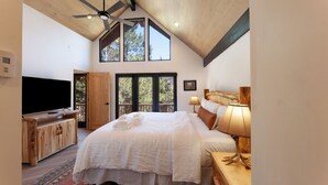 A spacious bedroom with a large bed, a television, and a wooden desk. There are large windows providing natural light and a view of trees.