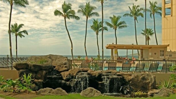 A pool area with a waterfall feature, surrounded by palm trees and a building with a balcony overlooking the pool. - Sugar Beach Resort  - Ocean View,   (Off Season Special Rates) (Kihei)
