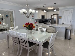 A modern kitchen with a glass dining table, white chairs, a chandelier, and stainless steel appliances.
