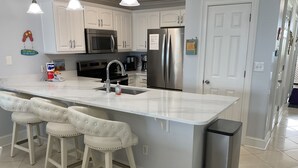 Una cocina con armarios blancos, electrodomésticos de acero inoxidable, una isla de mármol con taburetes y un bote de basura.