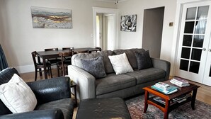 A living room with a gray sofa, black leather armchair, and ottoman, a coffee table with books, and a dining area with a wooden table and chairs. - Sunny Family Friendly 2 Bd1 Ba Sleeps 6, Safe, Fun & Walkable Grand Lake Oakland (Oakland)
