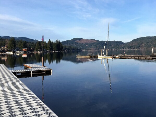 A calm lake with a dock, a sailboat, and a flagpole with a flag. - Lakefront Paradise: Private Beach, Water Sports, Stunning Sunsets, Sleeps 8 (Bellingham)