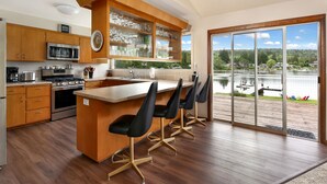 A modern kitchen with wooden cabinets, a central island with bar stools, a large window with a lake view, and a sliding glass door leading to an outdoor deck. - Lakefront Paradise: Private Beach, Water Sports, Stunning Sunsets, Sleeps 8 (Bellingham)