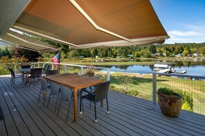 A wooden deck with a table and chairs, a flag, and a view of a lake and houses in the background. - Lakefront Paradise: Private Beach, Water Sports, Stunning Sunsets, Sleeps 8 (Bellingham)