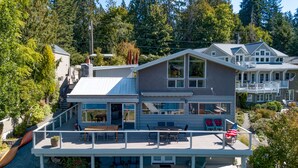 A two-story house with a covered patio, a large deck, and a view of the surrounding forest. - Lakefront Paradise: Private Beach, Water Sports, Stunning Sunsets, Sleeps 8 (Bellingham)
