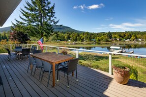 A wooden deck with outdoor furniture, a flag, and a view of a lake and mountains. - Lakefront Paradise: Private Beach, Water Sports, Stunning Sunsets, Sleeps 8 (Bellingham)
