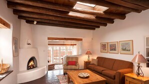 A cozy living room with a fireplace, wooden beams, a sofa, a coffee table, and a patterned rug. - Authentic Santa Fe Home~ Spacious, Vigas, Kiva Fireplaces and Walk to the Plaza (Santa Fe)