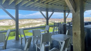 A covered outdoor seating area with wooden tables and chairs, overlooking a beach and a boardwalk.