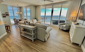 A spacious living area with a large window overlooking the ocean, a ceiling fan, and a dining area with a table and chairs.