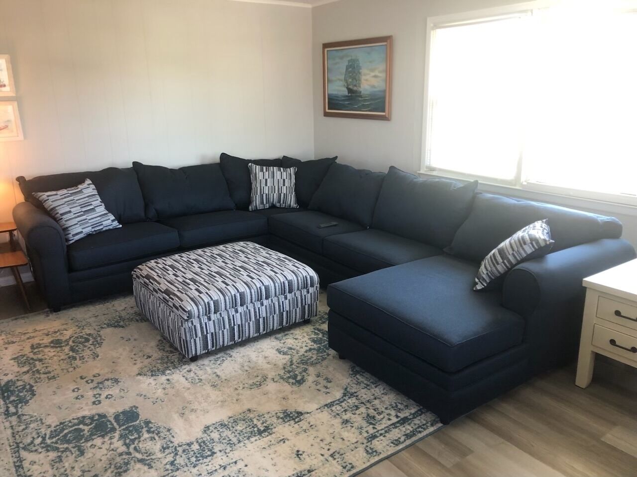 Una sala de estar con un sofá modular oscuro, un reposapiés estampado y una alfombra de colores claros.