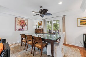 A dining area with a glass table, wooden chairs, a ceiling fan, and artwork on the walls.
