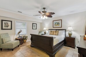 A bedroom with a bed, bedside tables, a chair, a desk, a ceiling fan, and artwork on the walls.