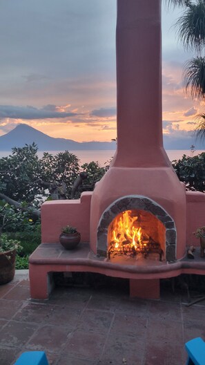 A stone fireplace with a fire burning inside, set in a red structure with a view of a mountain and sunset in the background.