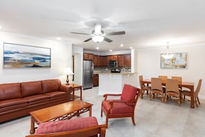 A spacious living room with a brown leather sofa, a wooden coffee table, and a red armchair. A dining area with a wooden table and chairs is adjacent to the kitchen, which features wooden cabinets and a stainless steel refrigerator.