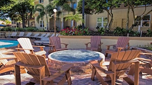 A poolside area with Adirondack chairs arranged around a hot tub, a pool, and a building with balconies in the background.