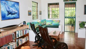 A cozy living room with a green sofa, wooden rocking chair, and a hammock. There is a bookshelf filled with books and a painting of a whale on the wall.
