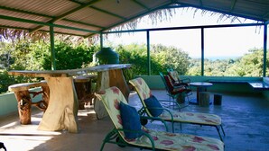 A covered outdoor seating area with a table and chairs, surrounded by lush greenery and a view of the landscape.