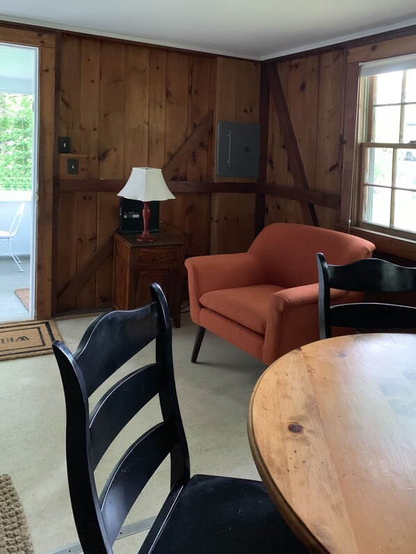 A room with wooden walls, a round wooden table, two black chairs, a red armchair, a small wooden side table with a lamp, and a window with a view of greenery. - Cape Cod Pond Cottage (Eastham)