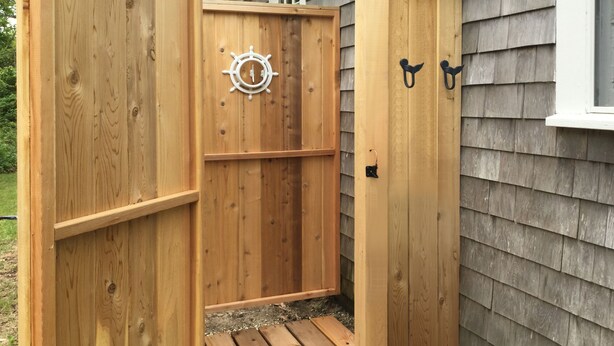 A wooden outdoor shower with a ship's wheel decoration, a small wooden platform, and two black hooks on the wall. - Cape Cod Pond Cottage (Eastham)