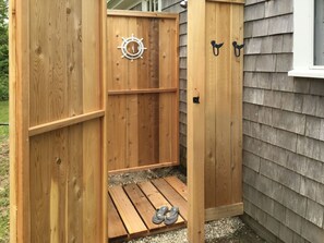Una ducha exterior de madera con un adorno de timón, una pequeña plataforma de madera y dos ganchos negros en la pared.