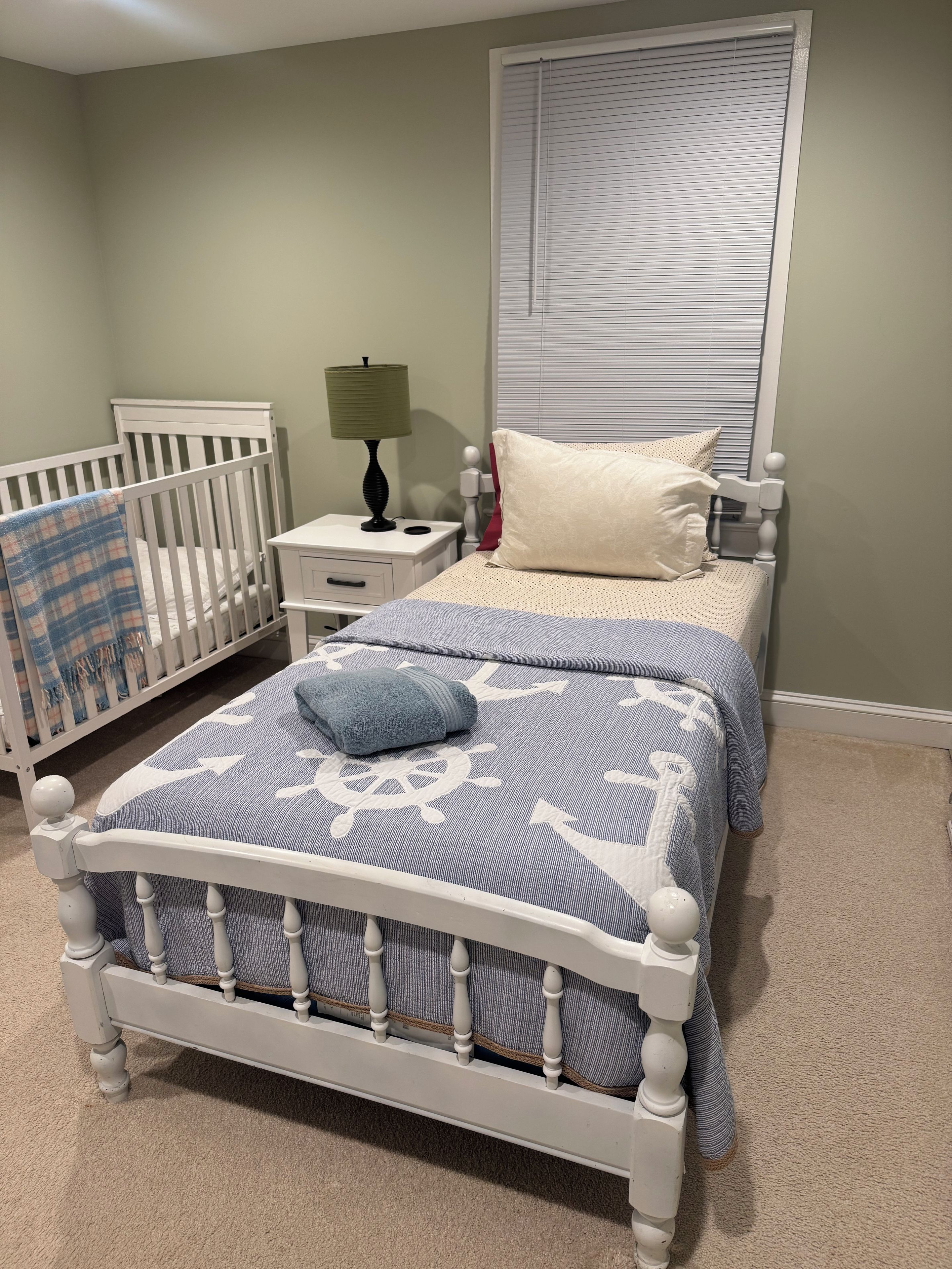 Une chambre à coucher avec un berceau, un lit recouvert d’une couvre-lit à motifs, une table de chevet avec une lampe et une fenêtre avec des stores.