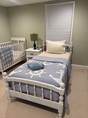 Un dormitorio con una cuna, una cama con colcha estampada, una mesita de noche con lámpara y una ventana con persianas.