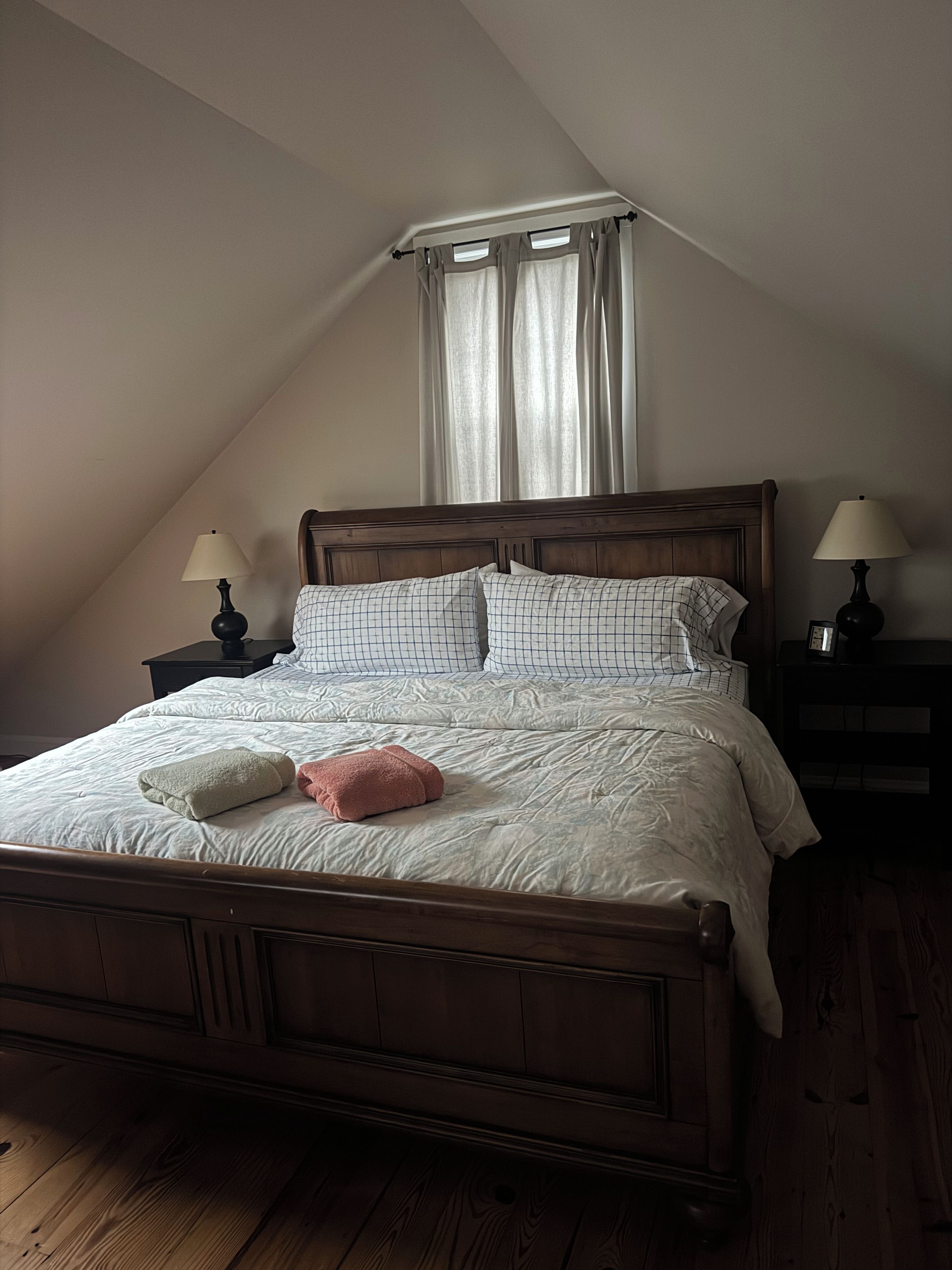 A bedroom with a wooden bed, two bedside tables with lamps, a window with curtains, and a sloped ceiling.