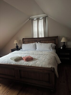 A bedroom with a wooden bed, two bedside tables with lamps, a window with curtains, and a sloped ceiling. - Fabulous Waterfront Property in the Heart of Wine Country (Peconic)
