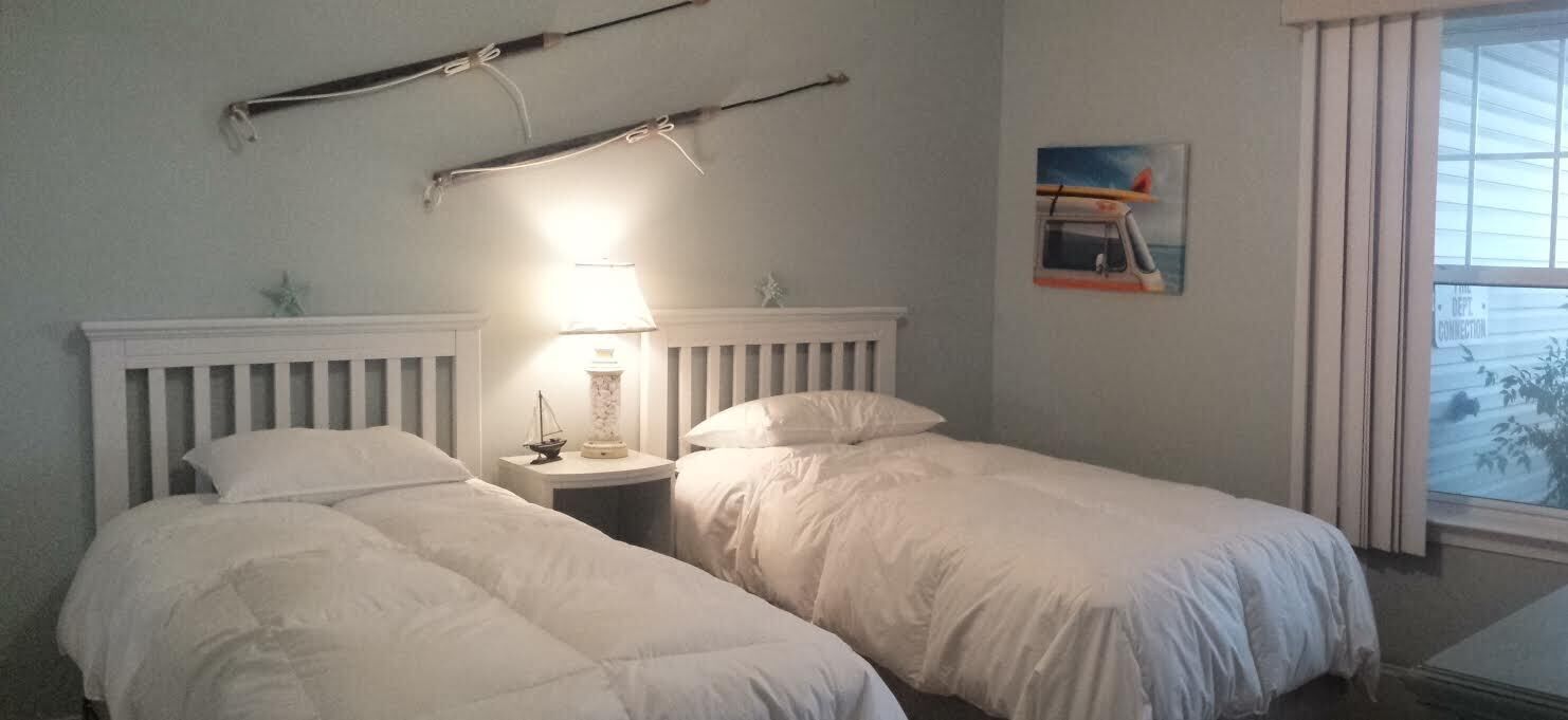 Une chambre avec deux lits simples, une table de chevet avec une lampe, un tableau représentant un bateau accroché au mur et une fenêtre avec des rideaux.