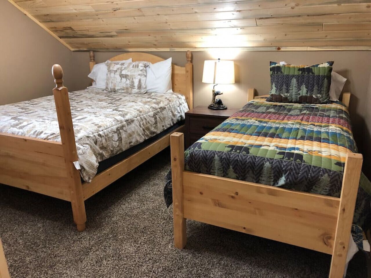 Dos camas de madera con ropa de cama diferente, una mesita de noche con lámpara y techo de madera.