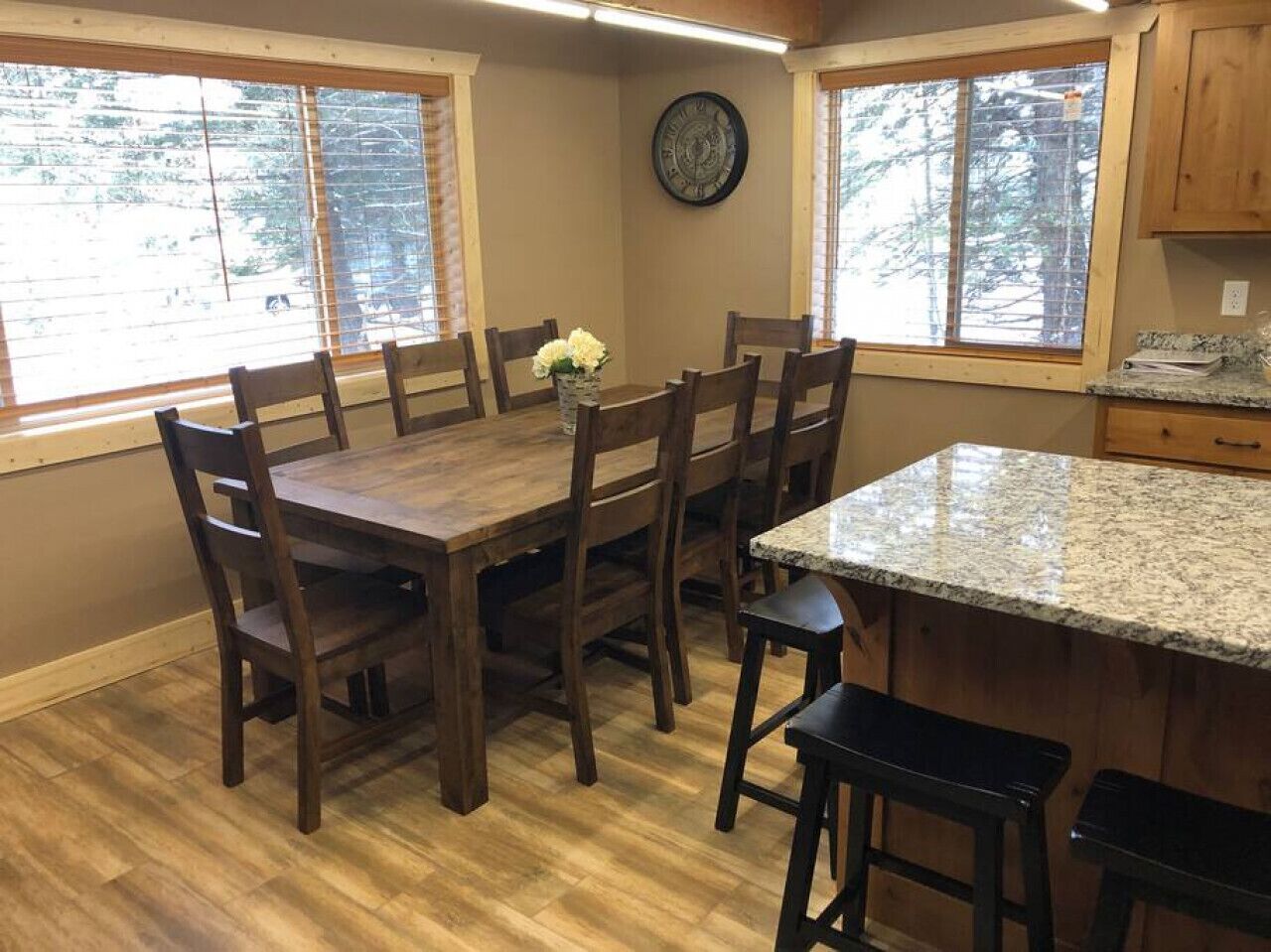 A kitchen with a wooden dining table, six chairs, a granite countertop, and a clock on the wall.