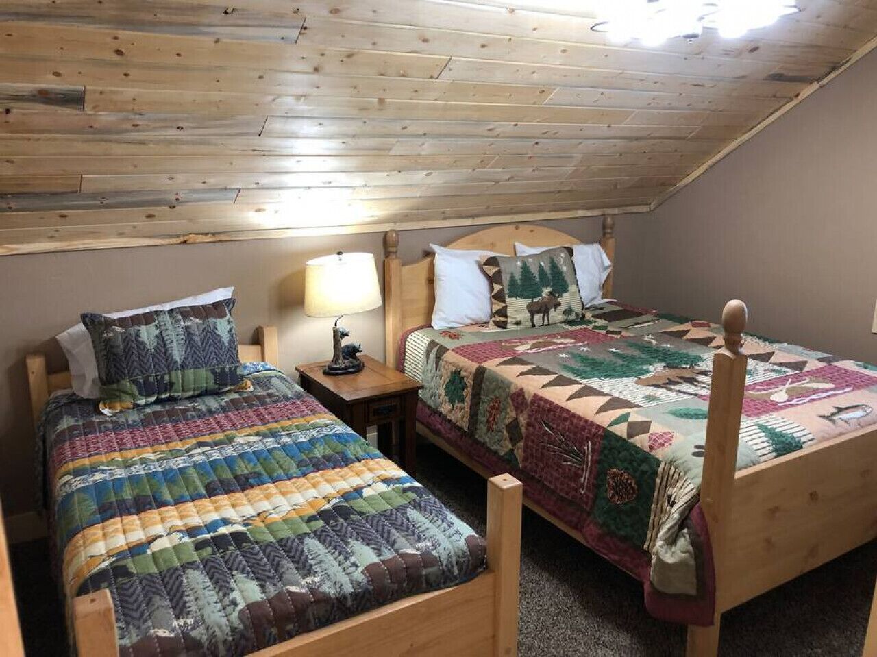 Une chambre mansardée chaleureuse avec deux lits en bois, chacun recouvert d’une courtepointe. Une table de chevet avec une lampe se trouve entre les deux lits.