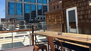 Una terrazza sul tetto con tavolo e sedie in legno, condizionatori d'aria e vista su un edificio moderno con finestre riflettenti.