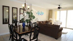 A dining area with a glass table, chairs, and a vase of flowers. There is a brown sofa and a lamp in the living area.