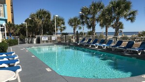 A pool area with blue lounge chairs, palm trees, and a lifeguard sign. - Luxurious and Elegant, 4BR Ocean Front, Get away! Ocean Blue - Myrtle Beach, SC! (Myrtle Beach)