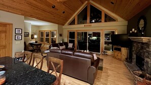 A spacious living room with a high ceiling, wooden beams, a fireplace, and a large window overlooking the interior.