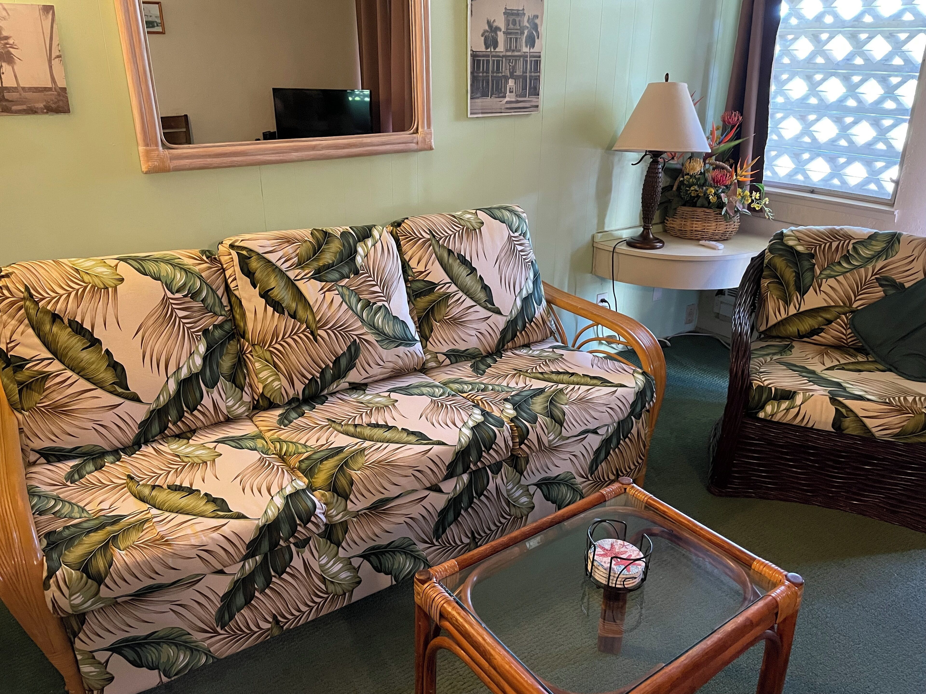 A living room with a floral-patterned sofa, a wicker armchair, a glass coffee table, a lamp, and a wall-mounted TV.